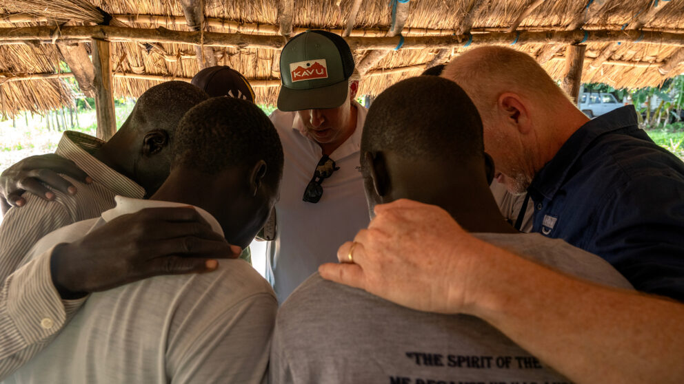 Pastors and visiting teammates praying together under a thatched shelter in Kajo Keji