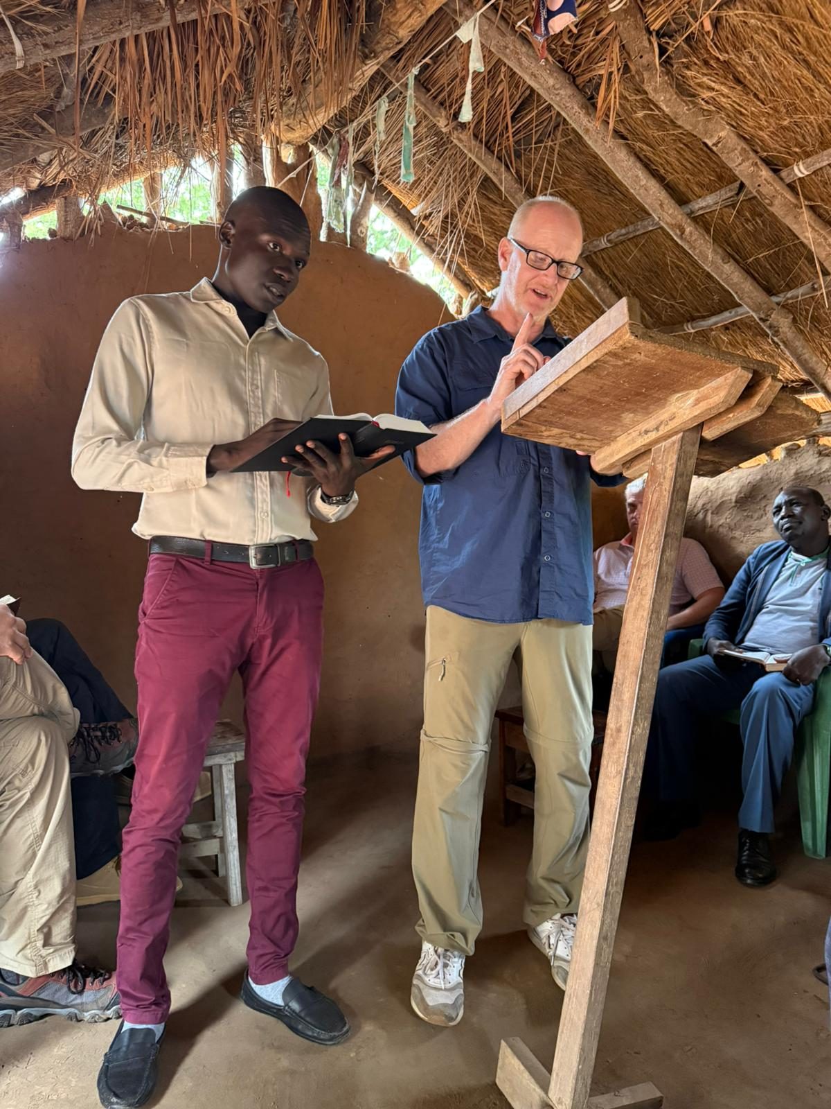 A visiting teammate and a South Sudanese leader teaching together from a wooden pulpit in a thatched church